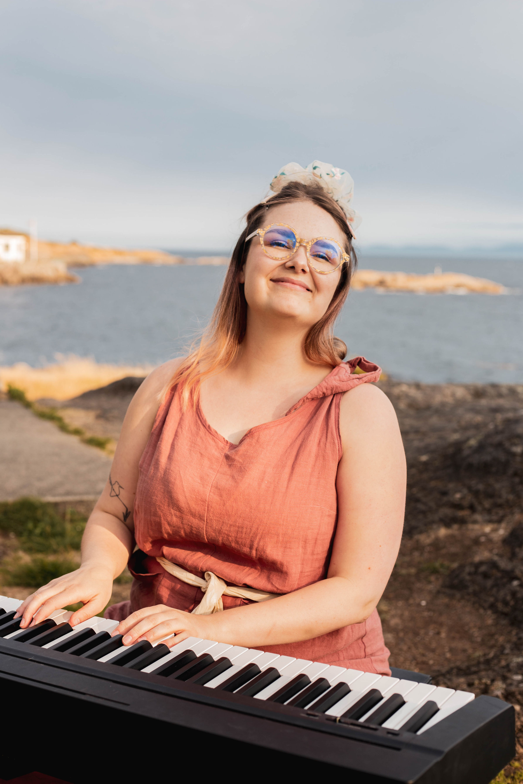 white non-binary person with short brown hair smiling with their eyes open wearing a pink toque, red button up and jean jacket, facing the camera standing in front of a tree.